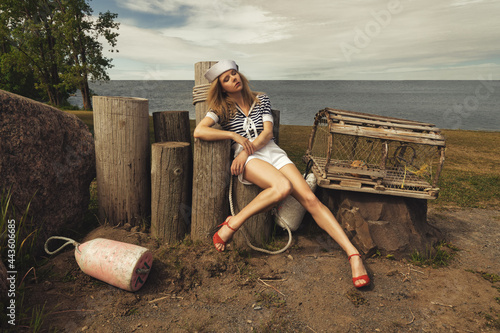 Young blonde woman with crimped hair, dressed in a sailor style outfit, sitting on wooden pillars in a lobster fisherman's cove photo