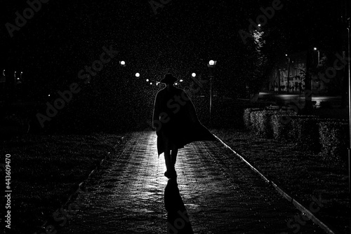 Canvas Print dramatic silhouette of a man in a hat and raincoat walking through the city at n