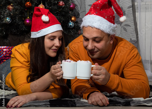 A man and a woman in red Christmas hats are holding a white mug with cocoa and marshmallows in their hands. Selective focus.