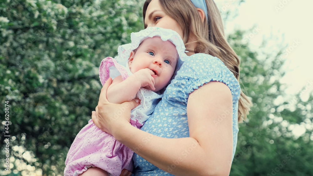 Careful blonde mother in blue dress holds in arms crying girl kid and lulls standing against blurred blooming fruit trees in park on spring day Careful blonde mother in blue dress holds in arms crying girl kid and lulls standing against blurred blooming fruit trees in park on spring day