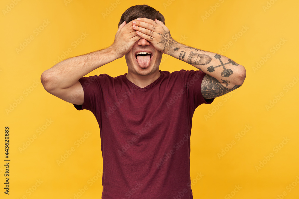 Naklejka premium Indoor shot of funny young male, closed his eyes with arms, shows tongue while waiting for surprise, isolated over yellow background