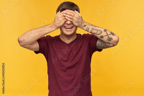Indoor shot of young male wears red t-shirt, smiles broadly and closed his eyes with arms while waiting for gift. Isolated over yellow background.