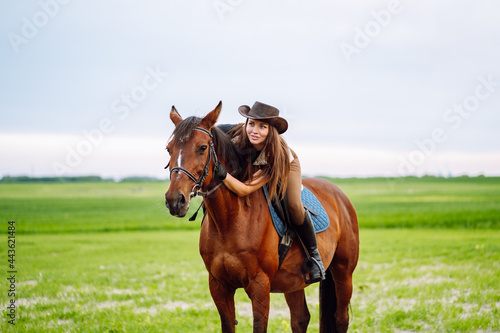 Young woman dressed in riding clothes and hat riding brown horse in green field © Olga