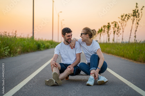 a man and a blonde are sitting on a long board on the road against the background of the sunset. A stylish and fashionable couple, happy faces, laughing