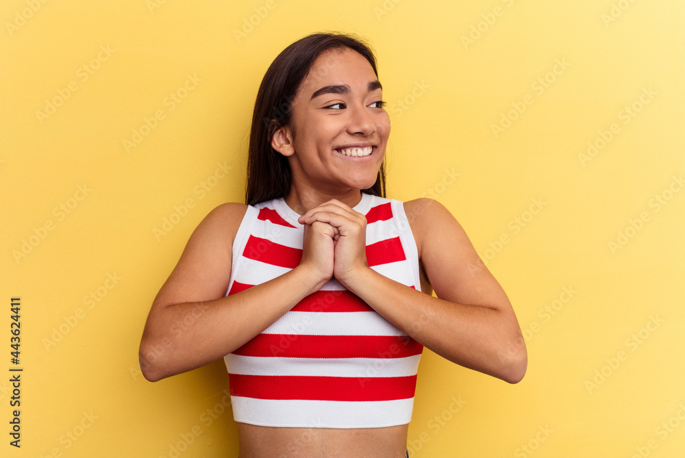Young mixed race woman isolated on yellow background keeps hands under chin, is looking happily aside. Young mixed race woman isolated on yellow background keeps hands under chin, is looking happily aside.