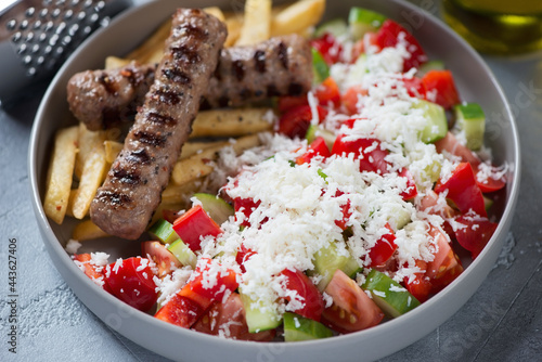 Foto Close-up of serbian shopska salad with grilled cevapi and fries in a grey plate,
