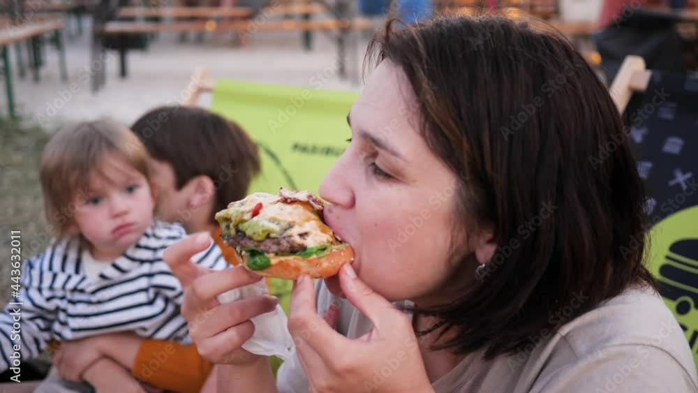Woman eating a tasty juicy part of a meat burger while her children watching her at street food trucks festival Woman eating a tasty juicy part of a meat burger while her children watching her at street food trucks festival