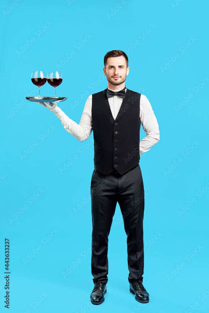 Obraz premium Full length portrait of elegant male waiter with glasses of wine over blue background