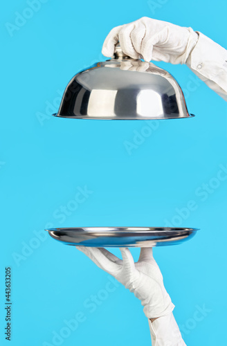 Elegant waiter's hands in white gloves holding silver tray and cloche on blue background photo