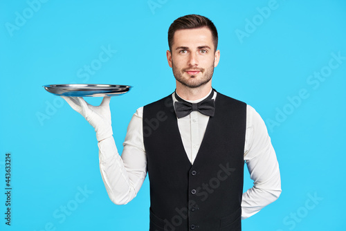 Attractive young waiter holding empty silver tray over blue background photo