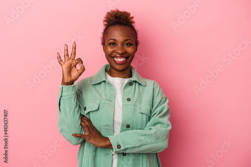 Young african american woman isolated on pink background winks an eye and holds an okay gesture with hand. © Asier