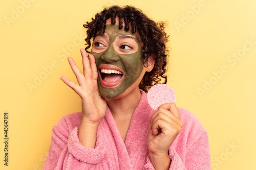 Young mixed race woman wearing a bathrobe holding a make-up remover sponge isolated on yellow background shouting and holding palm near opened mouth.