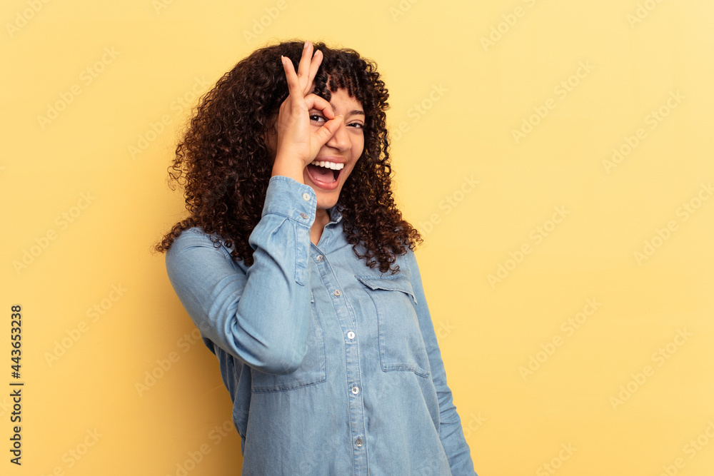 Young mixed race woman isolated on yellow background excited keeping ok gesture on eye. Young mixed race woman isolated on yellow background excited keeping ok gesture on eye.