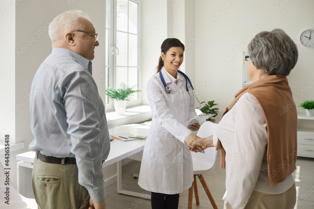 Obraz premium Female doctor shakes hands with an elderly woman who came to the clinic with her husband. Doctor welcomes or congratulates couple of patients on successful recovery. Healthcare concept for seniors.