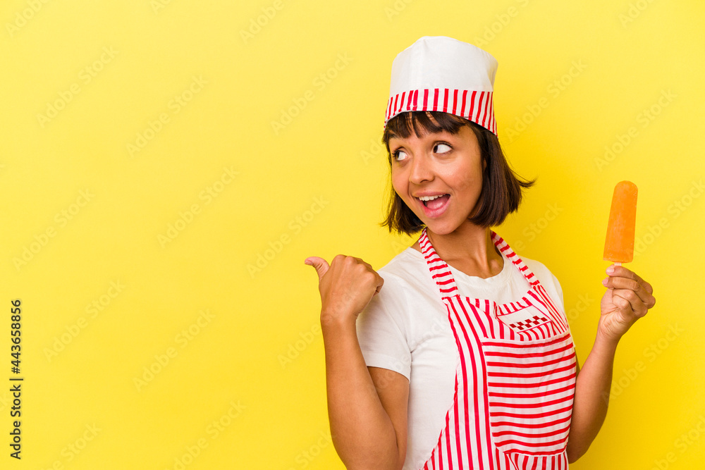 Young mixed race ice cream maker woman holding an ice cream isolated on yellow background points with thumb finger away, laughing and carefree. Young mixed race ice cream maker woman holding an ice cream isolated on yellow background points with thumb finger away, laughing and carefree.