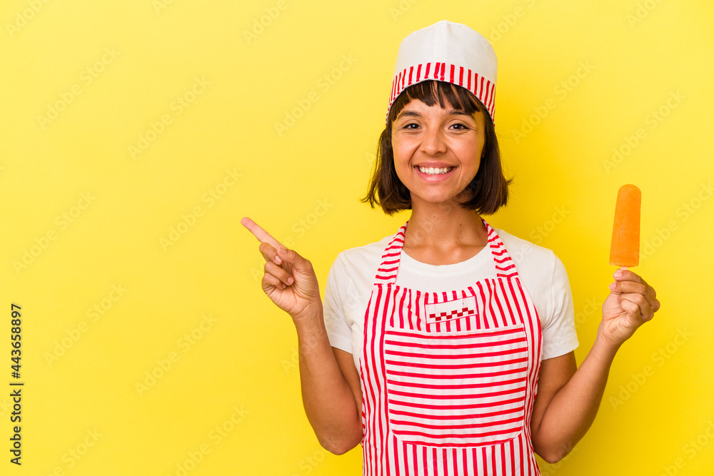 Young mixed race ice cream maker woman holding an ice cream isolated on yellow background smiling and pointing aside, showing something at blank space. Young mixed race ice cream maker woman holding an ice cream isolated on yellow background smiling and pointing aside, showing something at blank space.