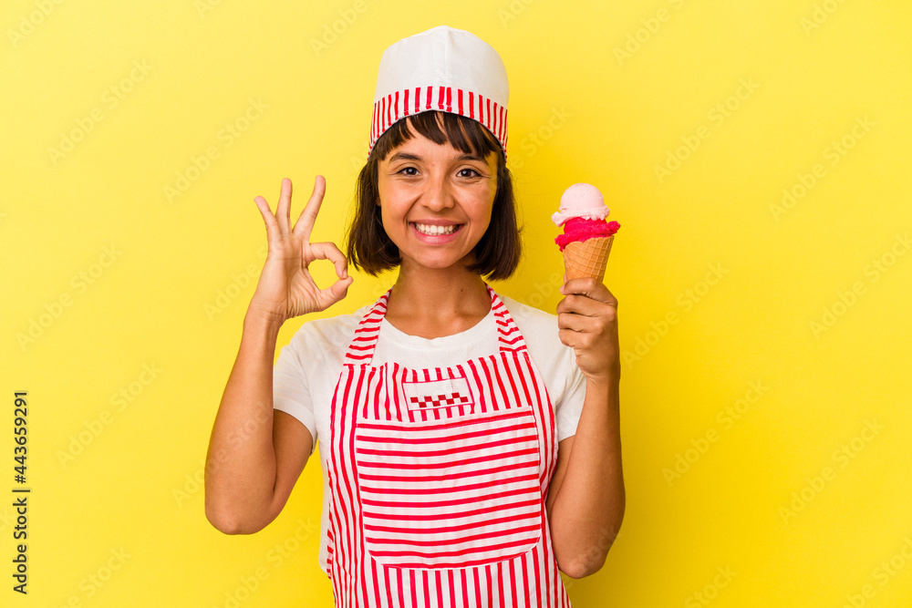 Young mixed race ice cream maker woman holding an ice cream isolated on yellow background cheerful and confident showing ok gesture. Young mixed race ice cream maker woman holding an ice cream isolated on yellow background cheerful and confident showing ok gesture.