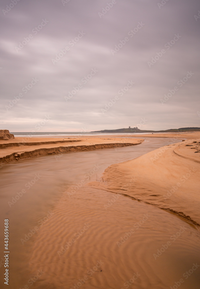 Dunstanburgh Castle in Northumberland, England, taken with the river meandering in the foreground & with cloudy skies overhead