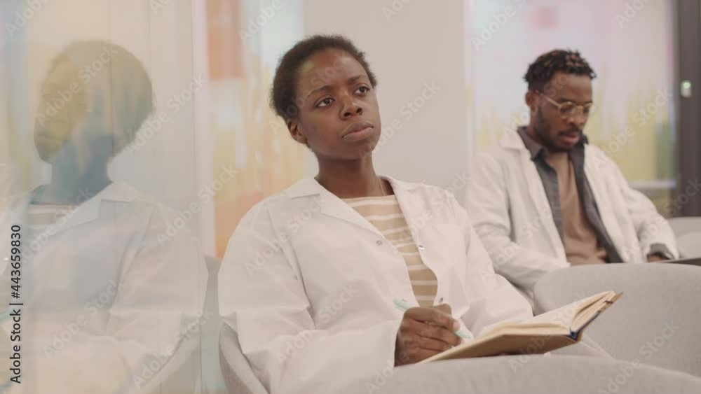 Medium shot of young African-American female medical student in white coat taking notes in copybook during lecture raising her hand to ask professor question Medium shot of young African-American female medical student in white coat taking notes in copybook during lecture raising her hand to ask professor question