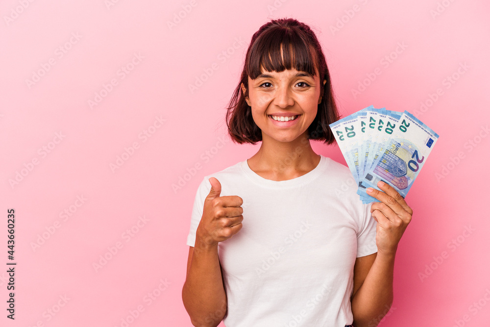Young mixed race woman holding bills isolated on pink background smiling and raising thumb up Young mixed race woman holding bills isolated on pink background smiling and raising thumb up