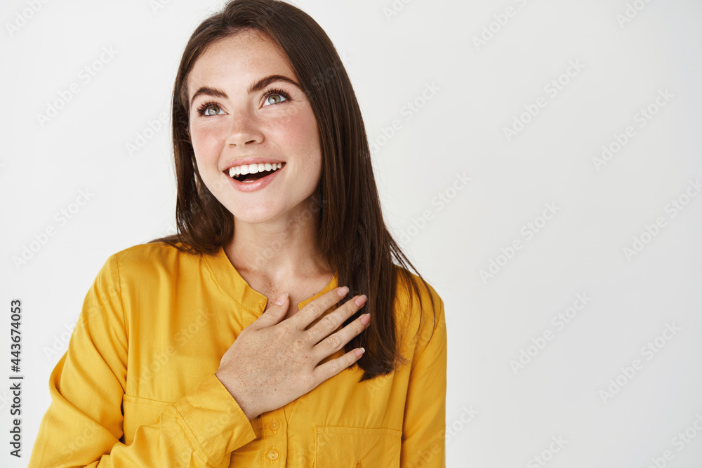 Close-up of dreamy and passionate woman holding hand on heart, looking with affection up and smiling, standing over white background Close-up of dreamy and passionate woman holding hand on heart, looking with affection up and smiling, standing over white background