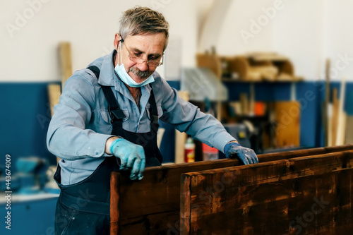 Obraz na plátně An elderly man paints wooden boards with dark varnish in a carpentry workshop