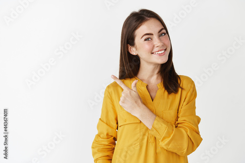 Happy young woman smiling, showing an advertisement, pointing at upper left corner and looking at camera, recommending product, standing over white background