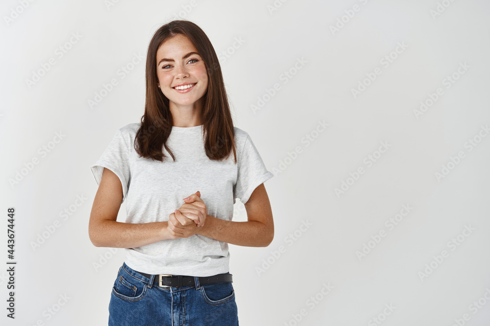 Young smiling woman consultant ready to help, looking friendly and holding hands together on chest, standing over white background Young smiling woman consultant ready to help, looking friendly and holding hands together on chest, standing over white background