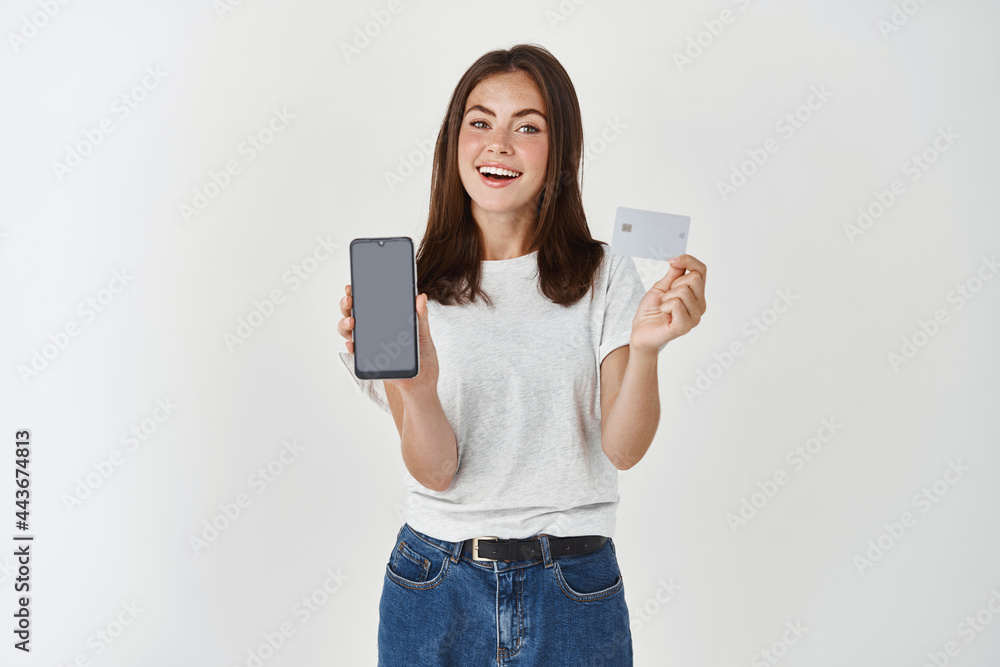 Portrait of a confident casual girl holding blank screen mobile phone and showing credit card isolated over white background Portrait of a confident casual girl holding blank screen mobile phone and showing credit card isolated over white background