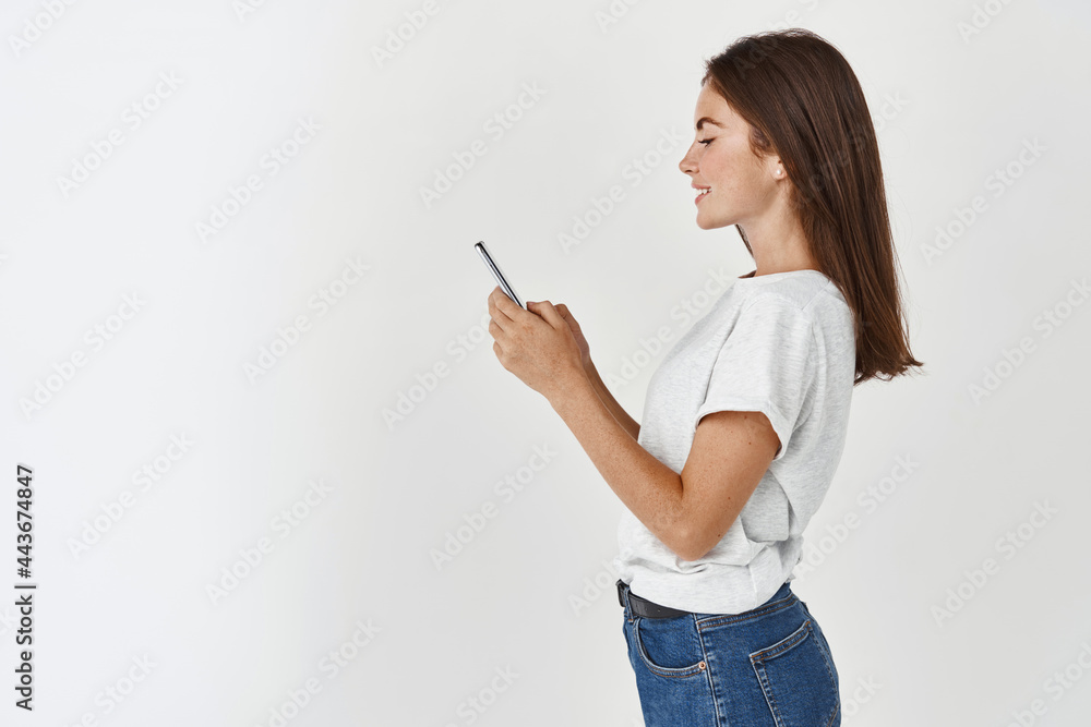 Fototapeta premium Profile portrait of beautiful brunette woman using mobile phone, reading message or browsing social media, standing happy against white background