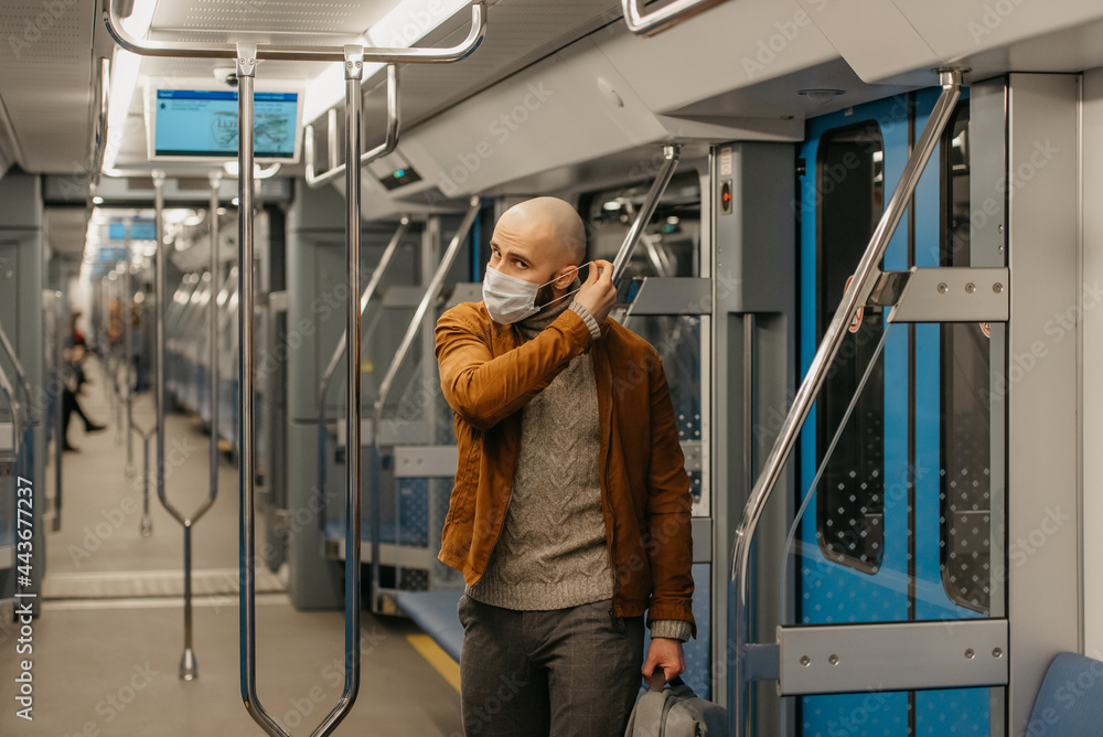 A man with a beard is putting on a medical face mask to avoid the spread of coronavirus in a subway car. A bald guy in a surgical mask against COVID-19 is keeping social distance on a train. A man with a beard is putting on a medical face mask to avoid the spread of coronavirus in a subway car. A bald guy in a surgical mask against COVID-19 is keeping social distance on a train.