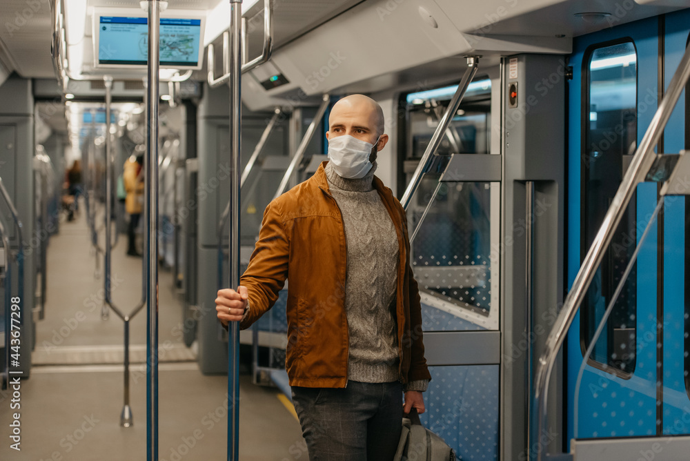 A man with a beard in a medical face mask to avoid the spread of coronavirus is riding and holding the handrail in a subway car. A bald guy in a surgical mask is keeping social distance on a train.