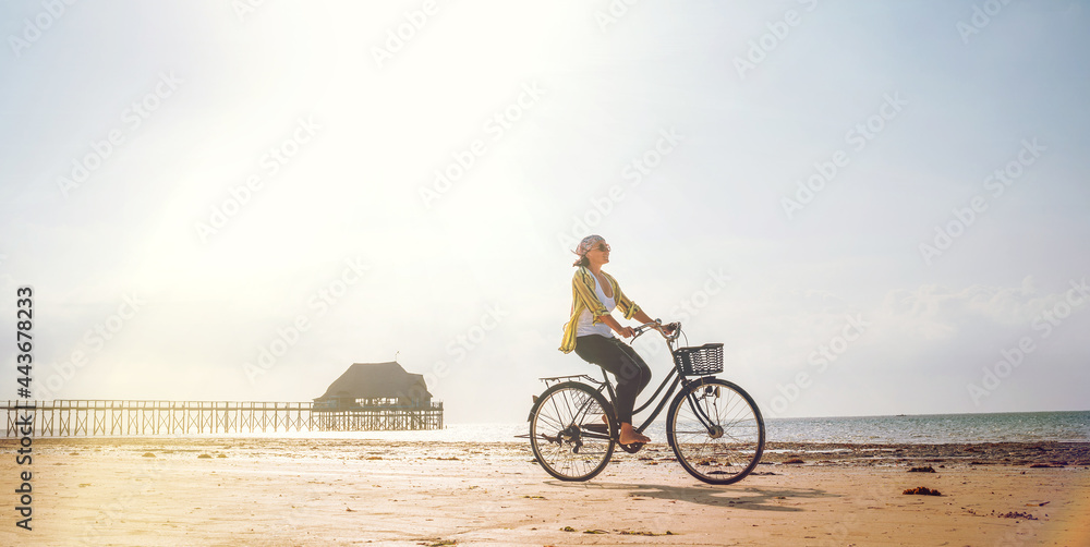 Fototapeta premium Young woman dressed light summer clothes riding old vintage bicycle with front basket on the lonely low tide ocean white sand coast on Kiwengwa beach on Zanzibar island, Tanzania.