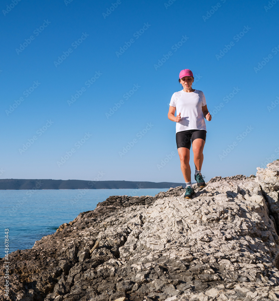 Young female dressed running sporty clothes and pink cap enjoying morning run race along the rocky calm sea coast. Sporty people trail running activities on vacation time concept image.