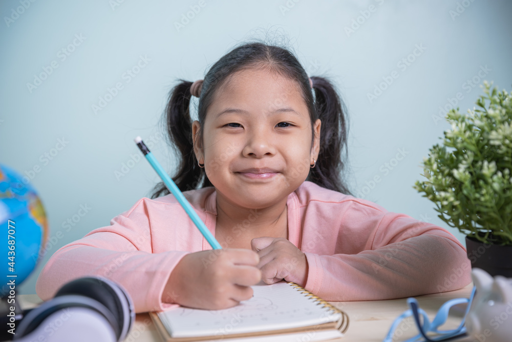 Student Asian kids girls smile happily looking at the camera writing on a notebook learning from home in the room personal. Concept learning online from a home social distance. Student Asian kids girls smile happily looking at the camera writing on a notebook learning from home in the room personal. Concept learning online from a home social distance.