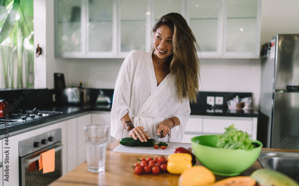 Joyful young Asian woman cutting cucumber and tomato and smiling during salad preparation