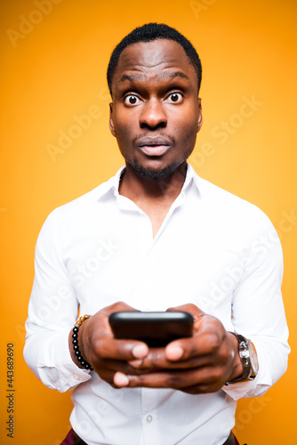 Handsome african american man holding a phone in his hand and is surprised by its power while standing on yellow background © DmitryStock