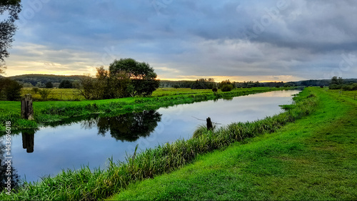 Gr  n blaue Abendd  mmerung mit schmalem orangenen Streifen am Horizont. Kanal Fluss  in dem sich die Landschaft und die dunklen Wolken spiegeln. Sumpfige Sumpf Landschaft.