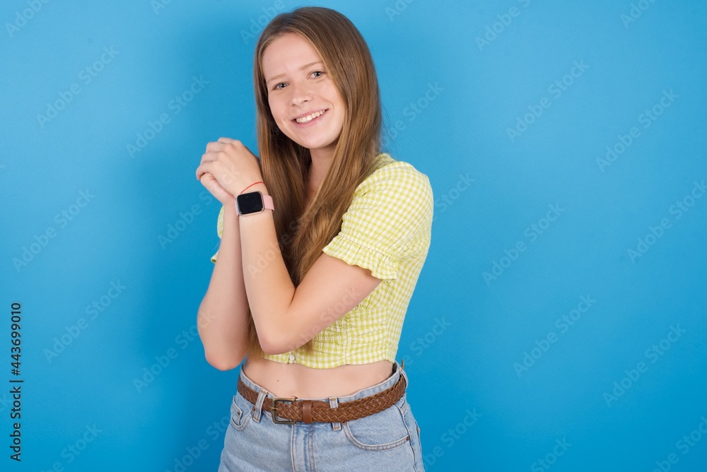 Dreamy young beautiful blonde woman standing against blue background with pleasant expression, closes eyes, keeps hands crossed near face, thinks about something pleasant