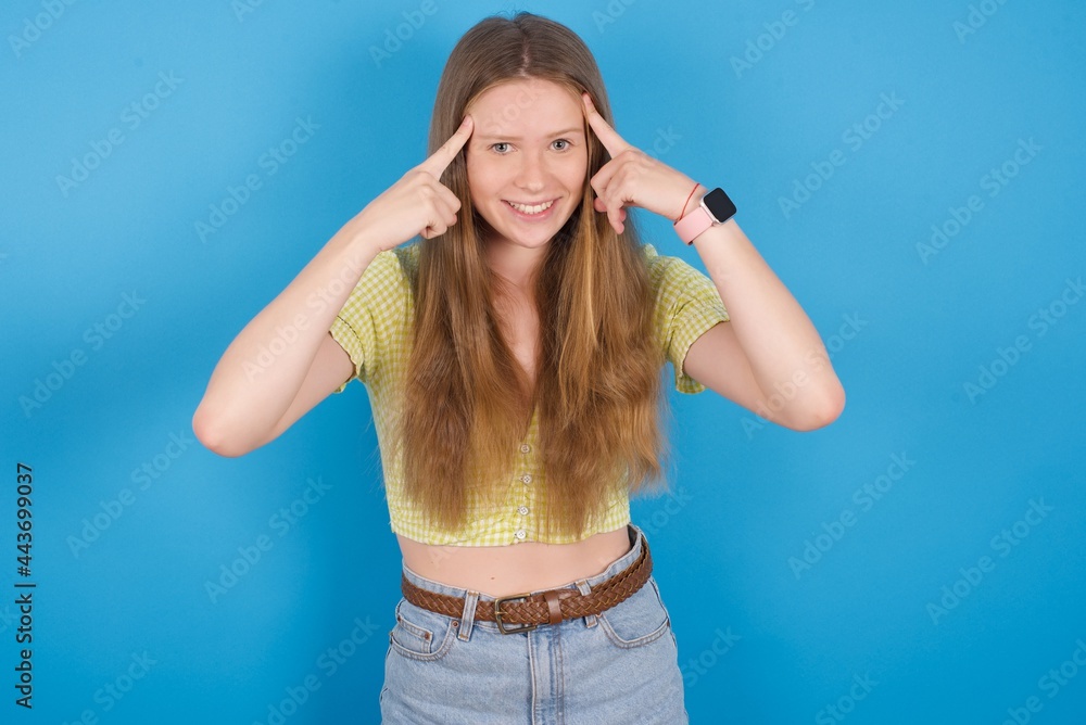 young beautiful blonde woman standing against blue background concentrating hard on an idea with a serious look, thinking with both index fingers pointing to forehead.
