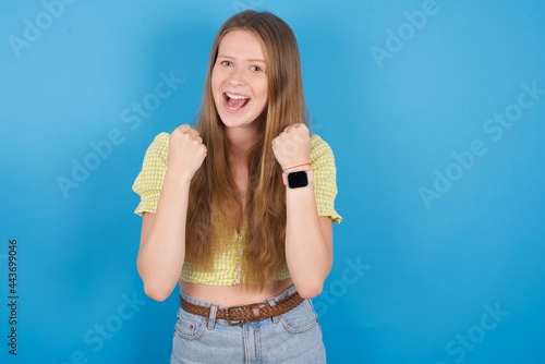 young beautiful blonde woman standing against blue background rejoicing success and victory clenching his fists with joy being happy to achieve her aim and goals. Positive emotions, feelings.