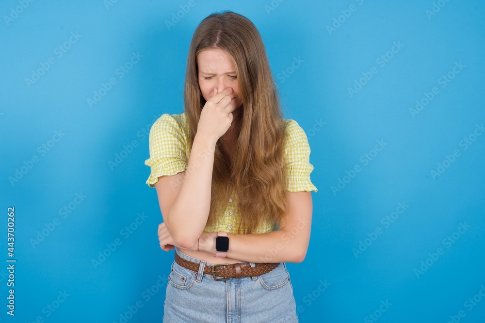 young beautiful blonde woman standing against blue background, holding his nose because of a bad smell. young beautiful blonde woman standing against blue background, holding his nose because of a bad smell.
