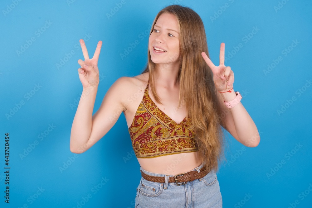 Isolated shot of cheerful young beautiful blonde woman standing against blue background makes peace or victory sign with both hands, feels cool. Isolated shot of cheerful young beautiful blonde woman standing against blue background makes peace or victory sign with both hands, feels cool.