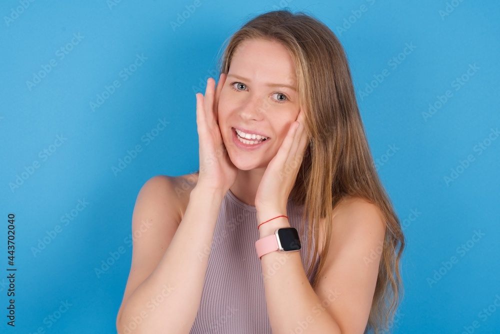 Happy young beautiful blonde woman standing against blue background touches both cheeks gently, has tender smile, shows white teeth, gazes positively straightly at camera, Happy young beautiful blonde woman standing against blue background touches both cheeks gently, has tender smile, shows white teeth, gazes positively straightly at camera,