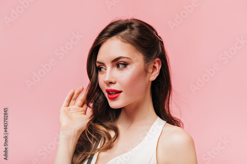 Lady in white top touches her wavy hair. Shot of brunette woman with red lips on pink background