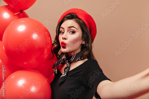 Curly girl in red beret and silk scarf whistles, holds balloons and makes selfie © Look!