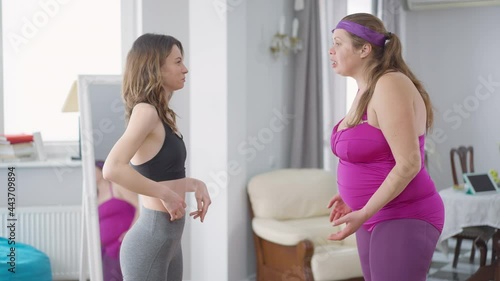 Side view portrait of obese and slim Caucasian women arguing indoors. Medium shot of angry overweight lady yelling as slender friend shouting. Conflict of two friends training at home photo
