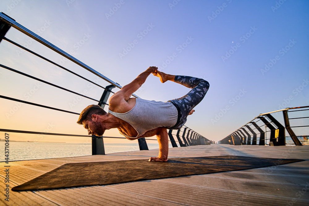 Fit man doing a one hand stand while practicing yoga alone near the ocean against sky at dusk or dawn