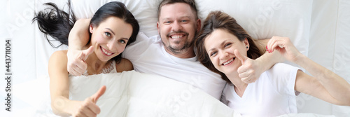 Obraz na plátně Two smiling women with man lie in bed and hold their thumbs up