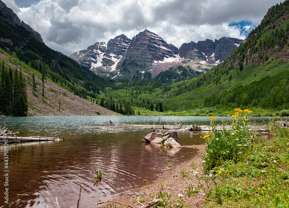 View of Maroon Bells in Aspen, Colorado. A mountain range with snow can be seen as well as clouds, a forest and valley, and a lake.